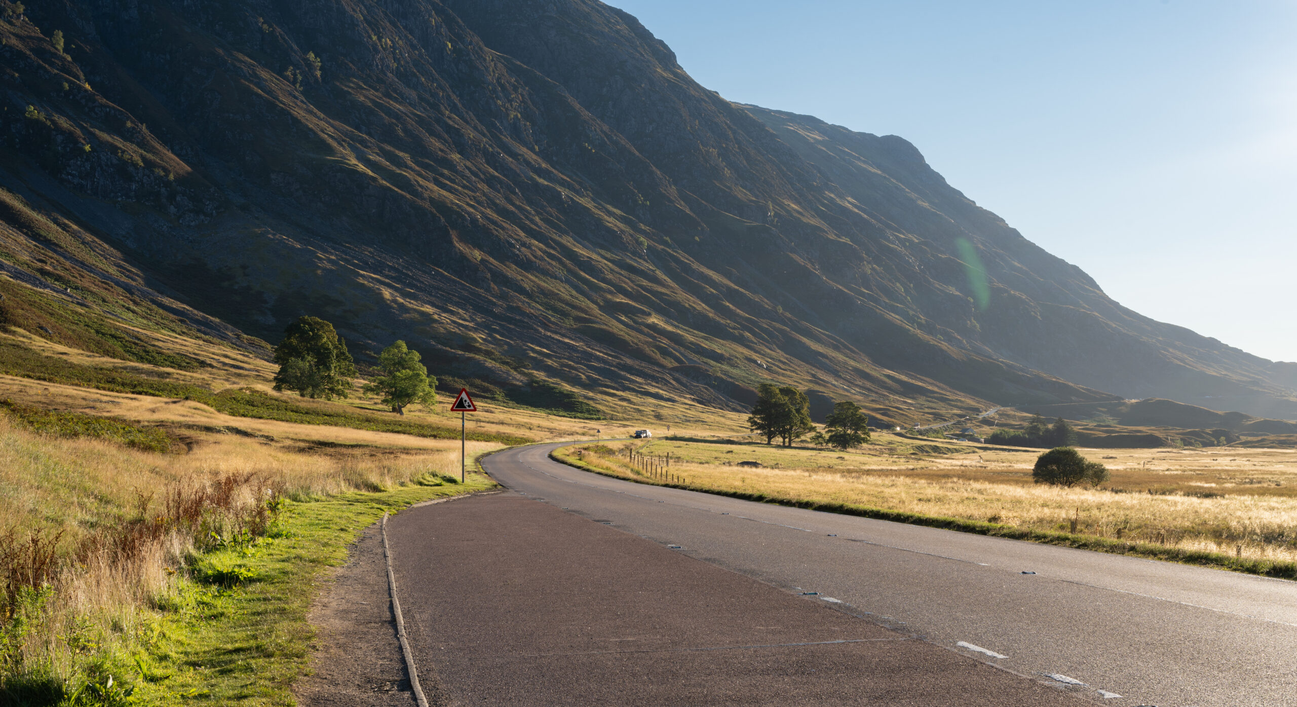 A quiet Highland road curving below a sunlit mountainside
